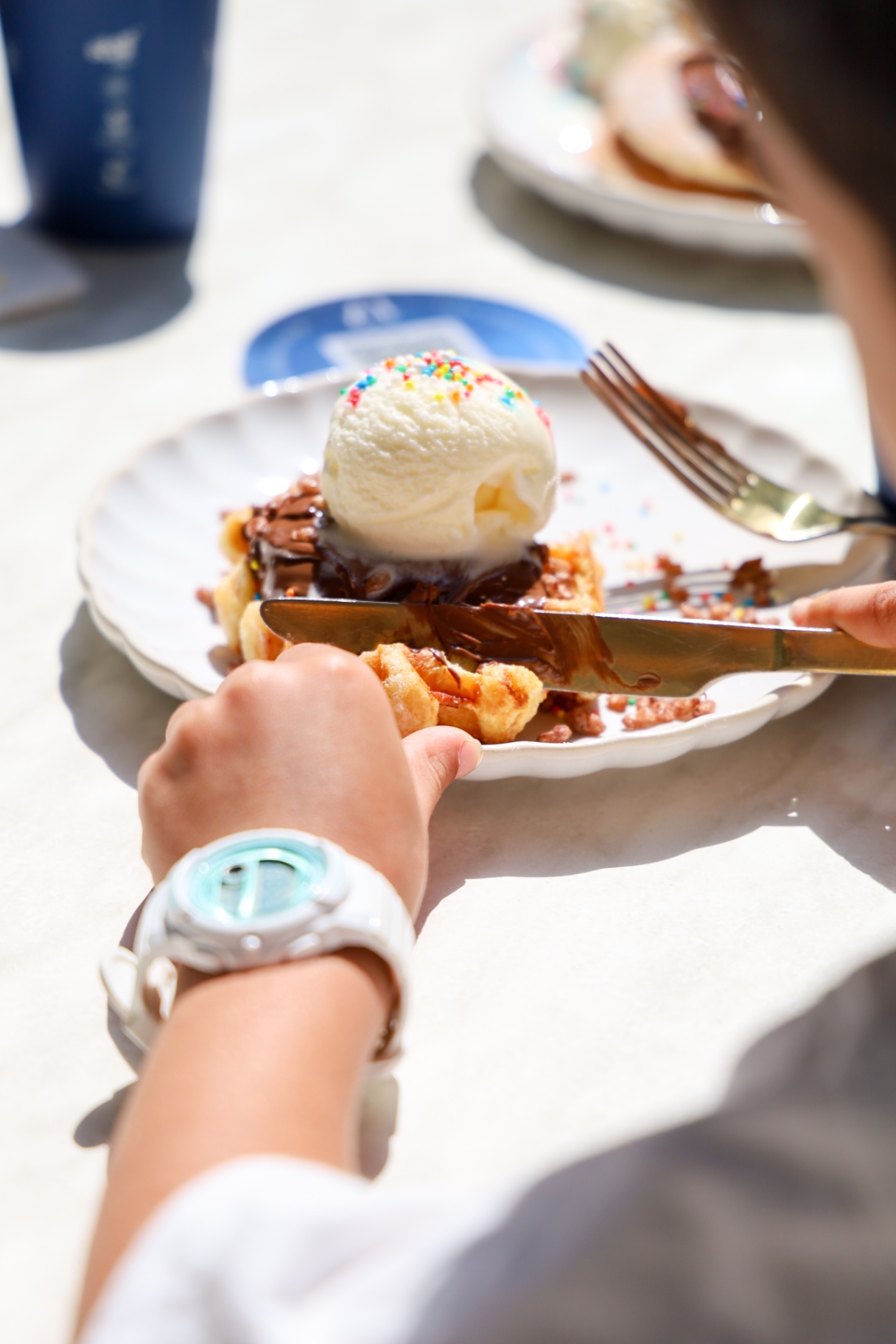 Waffle topped with chocolate sauce, sprinkles and a scoop of vanilla ice cream, a child cutting in